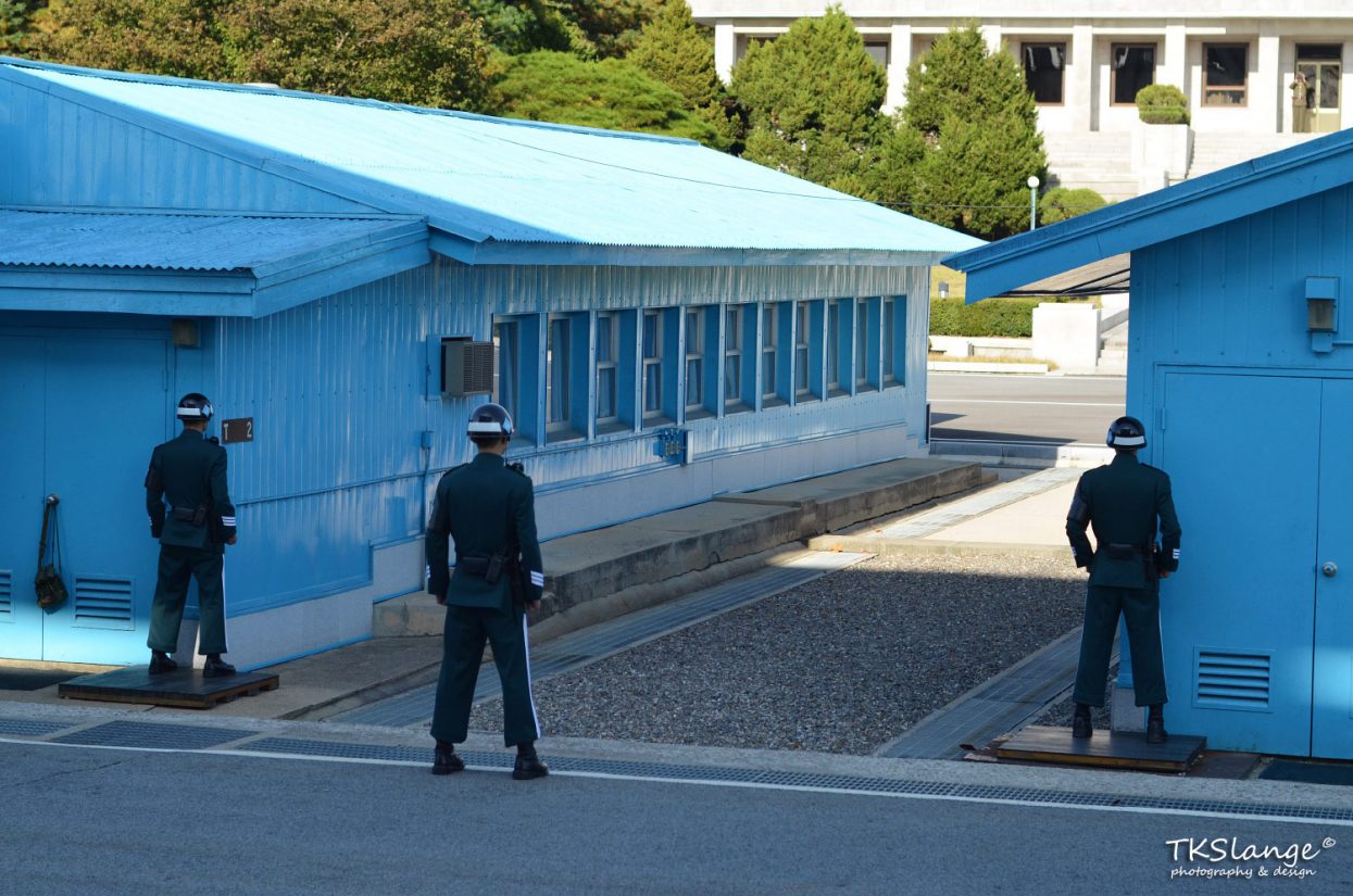 ROK soldiers facing North Korea. In the doorway of the building across stands a North Korean soldier with binoculars.